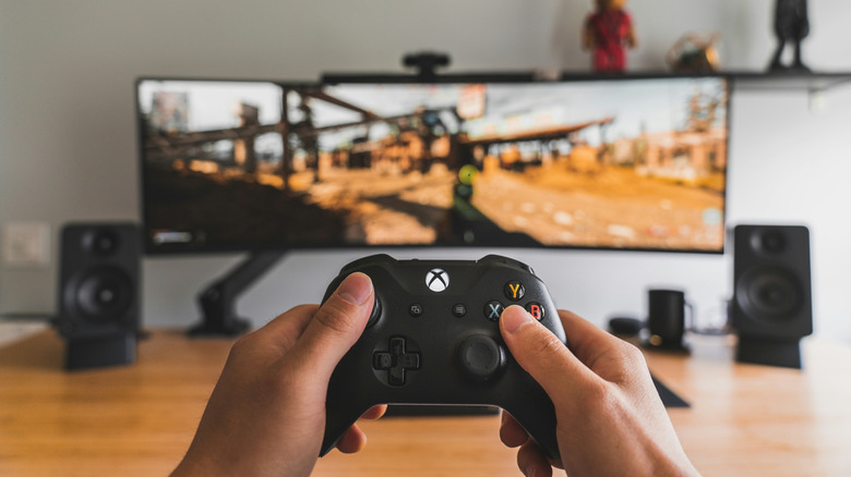 A person holds an Xbox controller as a game runs on a monitor in the background.