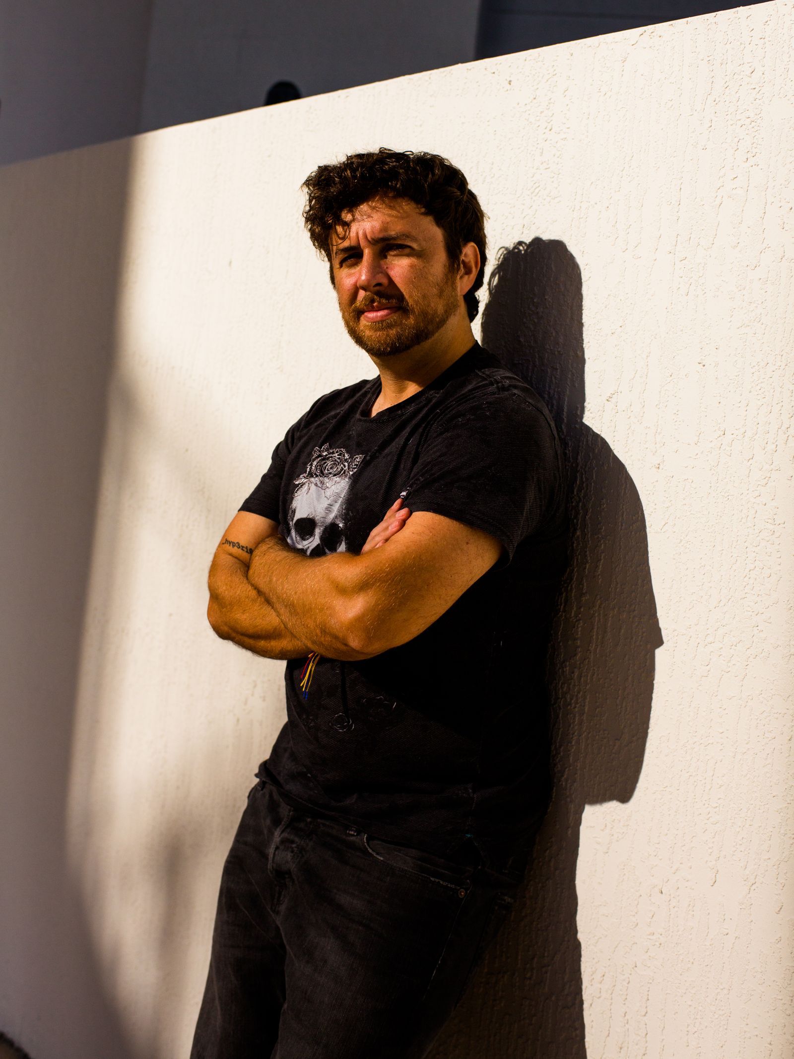 A person crossing their arms and leaning against a white wall in bright sunlight