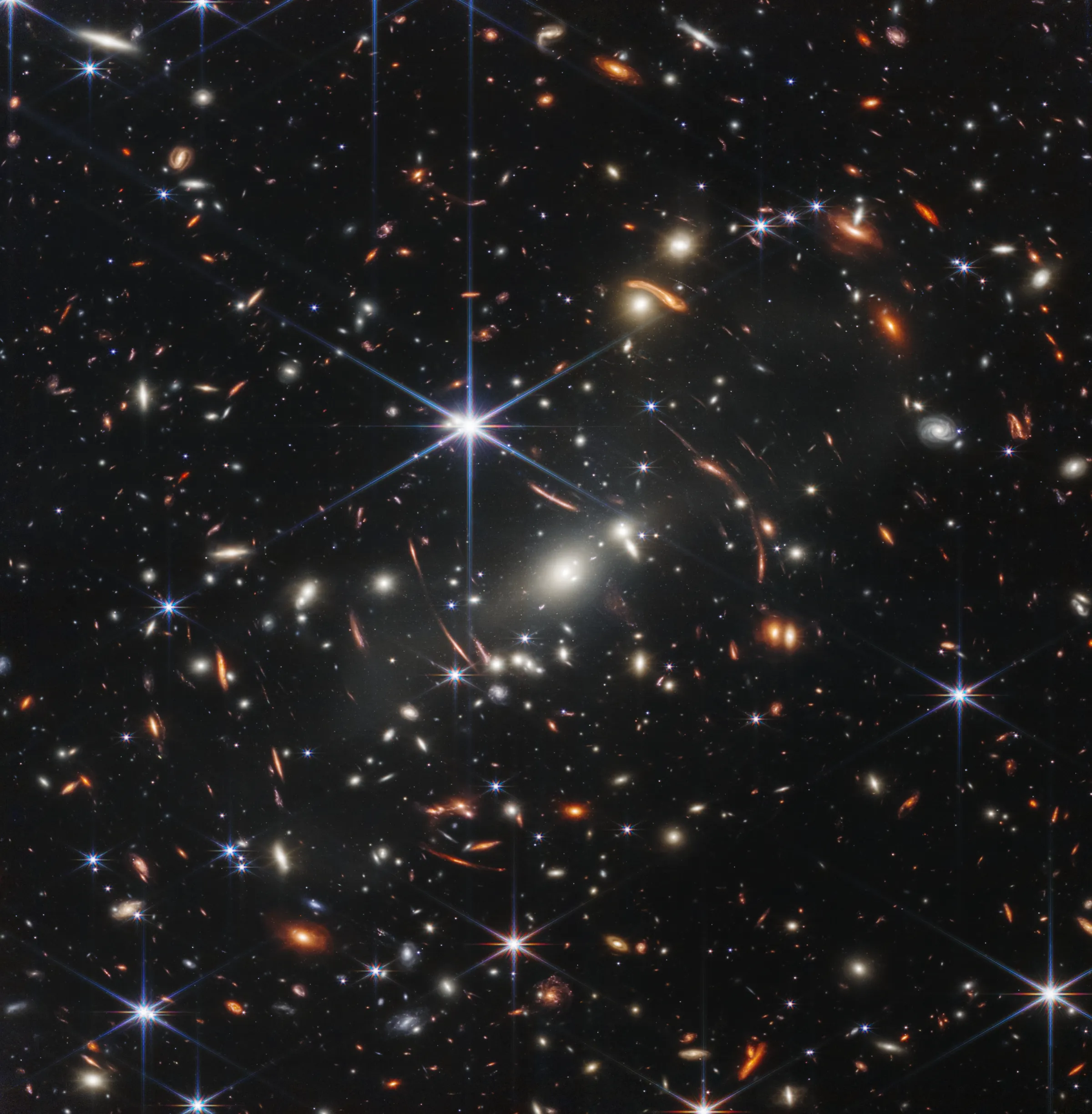 This image shows a spiral galaxy that is dominated by a bright central region. The galaxy has blue-purple hues with orange-red regions filled with stars. Also visible is large diffraction spike, which appears as a star pattern over the central region of the galaxy. Lots of stars and galaxies fill the background scene